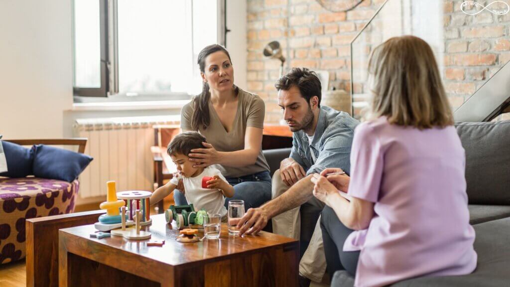A young child is playing with toys at a table with their mom and dad sitting on a couch behind them. The parents are talking to a therapist who has their back to the camera. The mom has her hands on the child's head.