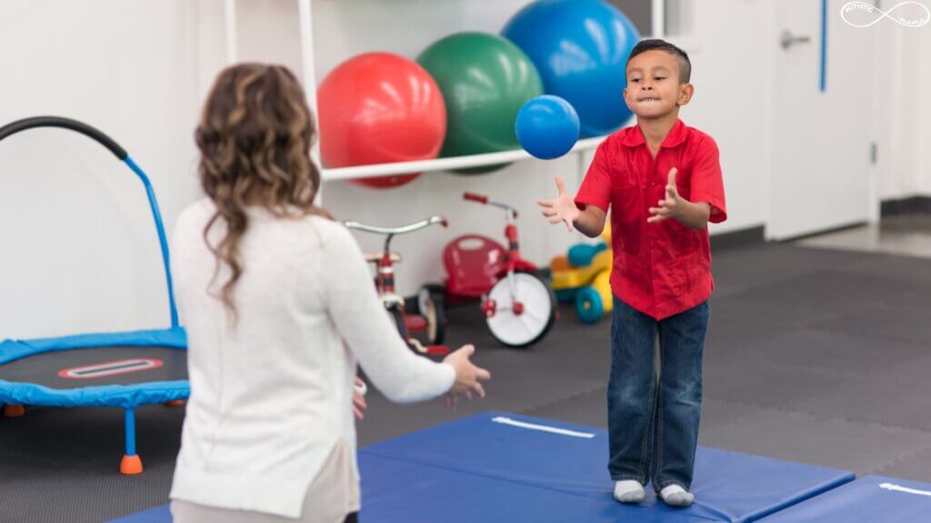 Young boy stands in a therapy clinic throwing a ball with a therapist who is kneeling and facing the boy.