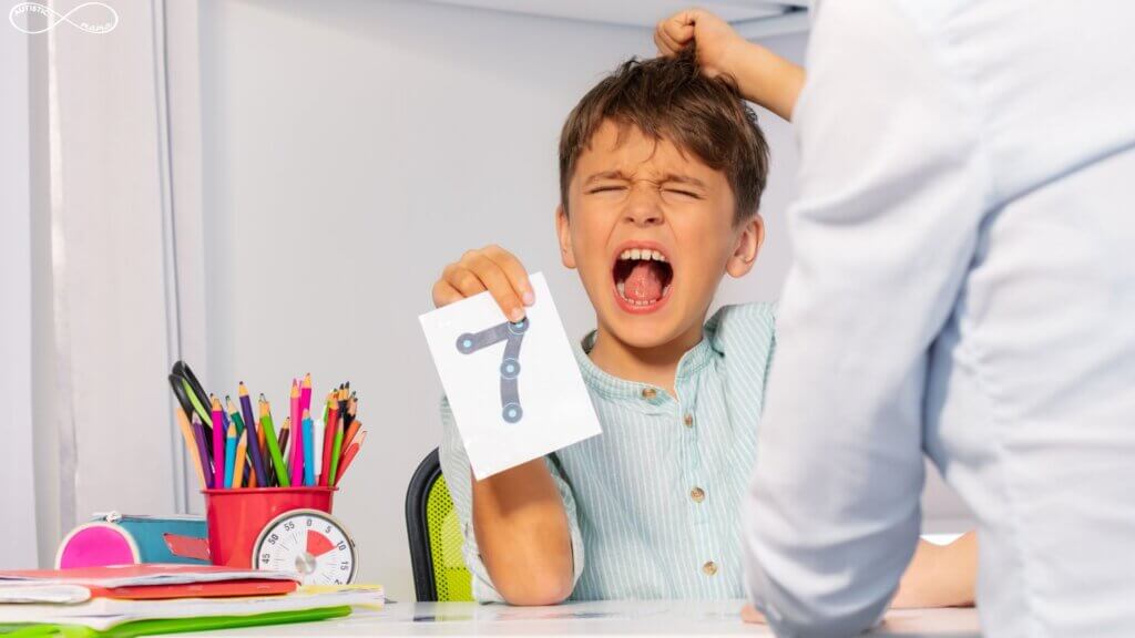 Young boy sits at a table with his therapist. He is pulling his hair and has an angry expression on his face. He is holding a card with the number seven on it.