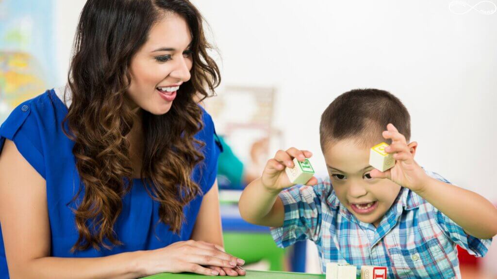A young boy is playing with blocks at a table and his therapist is sitting next to him. They're both smiling.